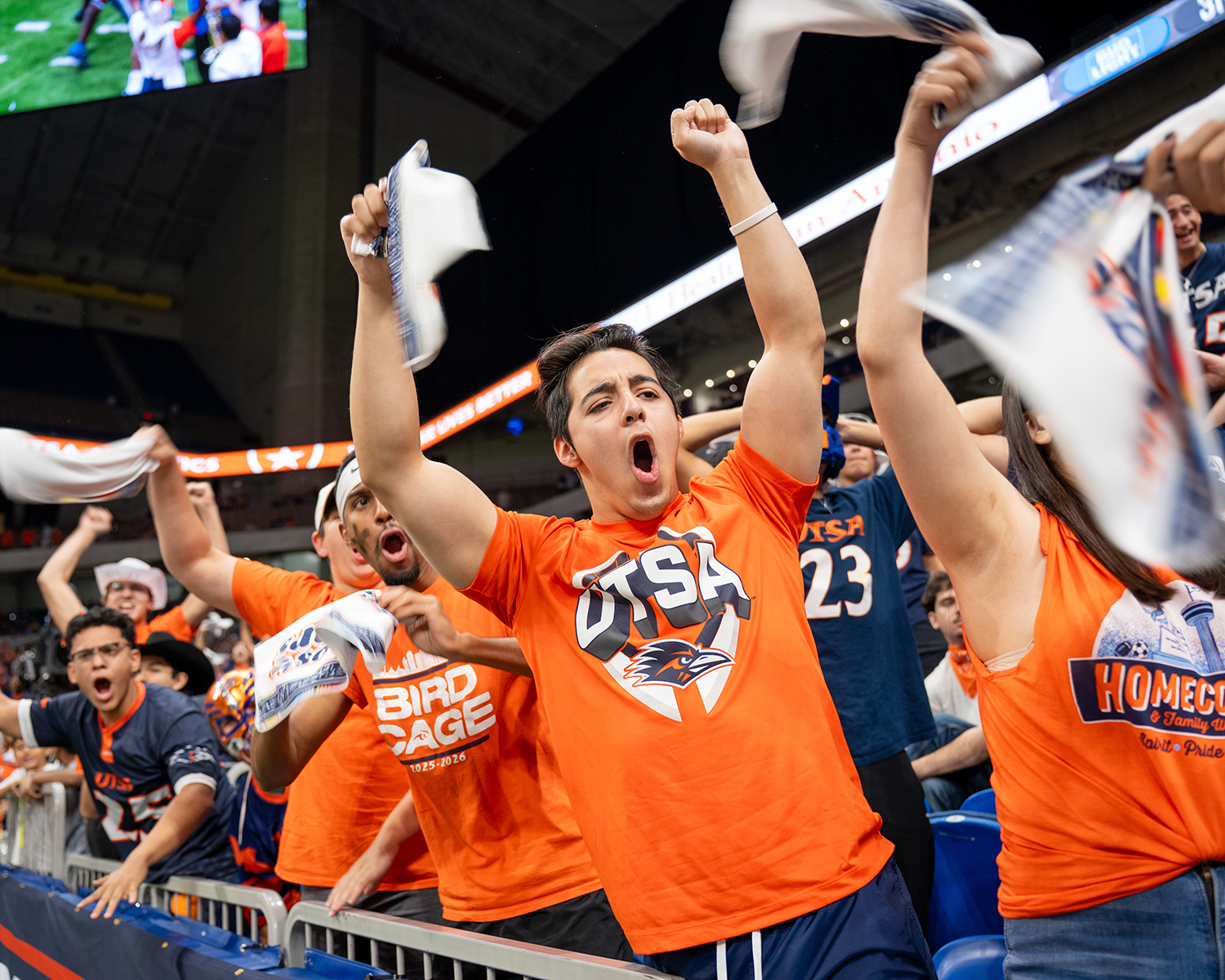 A crowd of students in the stands cheering wearing orange and blue school color t-shirts while spining towels over their heads.